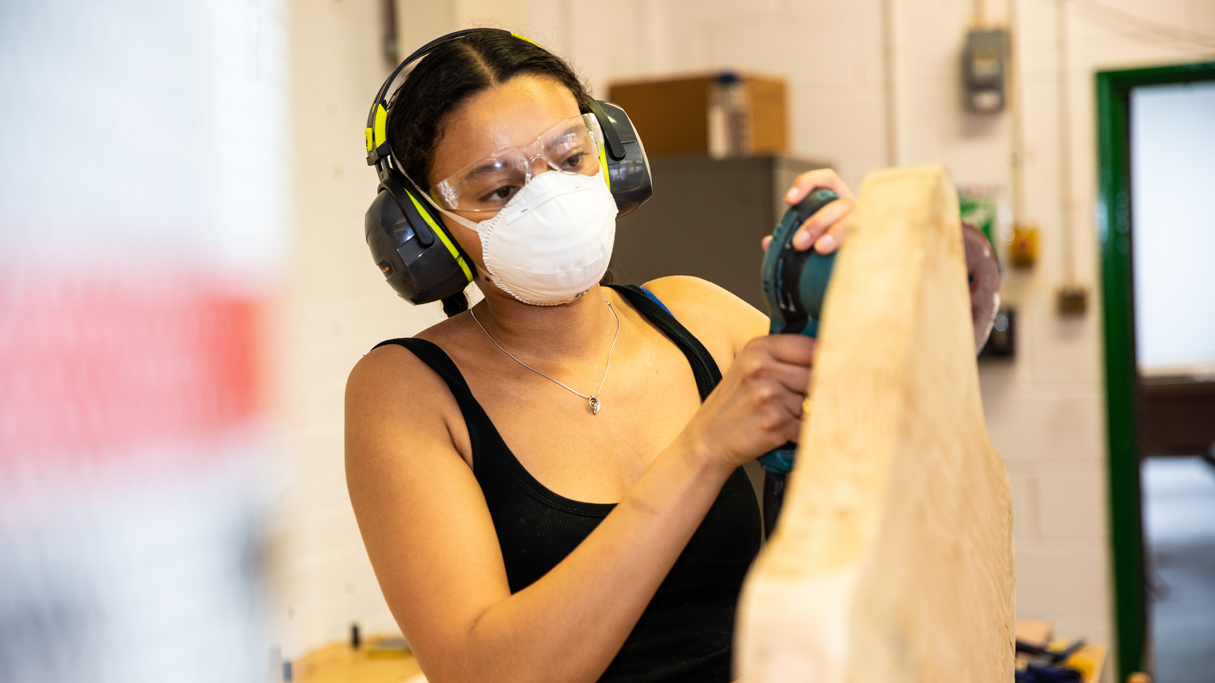 Female student in protective mask and ear defenders works on a wooden sculpture in the art studio