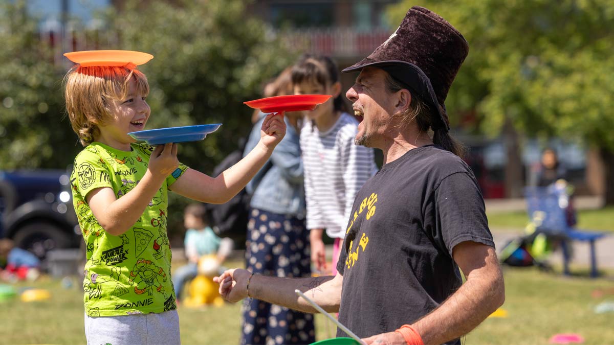 University of Reading's Community Festival 2025. A child laughing and smiling in the sun while spinning plastic plates with an adult in a large floppy hat.